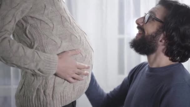 Side view of smiling Caucasian man in eyeglasses caressing his pregnant wifes belly and listening to babys heartbeat. Happy father waiting for his girlfriend to give birth. Pregnancy, gestation. — Stock Video