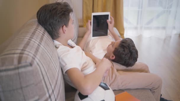 Vue latérale de la brune garçon caucasien parlant à son frère jumeau assis sur le canapé à la maison. Frères et sœurs se reposant ensemble après avoir joué au football . — Video