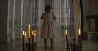 Mum ışığında duran tuhaf, beyaz bir kızın arka görüntüsü. Garip bir çocuk kameraya dönüp bebeği tutuyor. Korku, korku, tuhaflık..