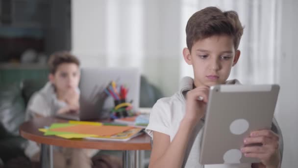 Πορτρέτο της νεαρής μελαχρινής Καυκάσιας αγόρι γυρίζει πίσω, κοιτάζοντας δίδυμο αδελφό του χρησιμοποιώντας φορητό υπολογιστή, και να κρατήσει για τη μελέτη. Schoolchild χρησιμοποιώντας δισκίο για την e-learning. Σύγχρονες τεχνολογίες, εκπαίδευση. — Αρχείο Βίντεο