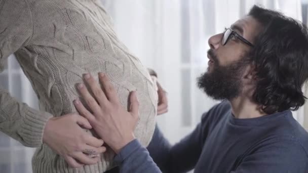 Happy Caucasian man listening to baby hitting and kissing his pregnant wifes belly. Bearded guy in eyeglasses waiting for his girlfriend to give birth. Pregnancy, gestation. — Stock Video