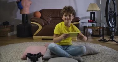 Evde yerde oturmuş video oyunları oynayan gülümseyen beyaz gencin portresi. Akıllı telefon kullanıp ekrana bakan mutlu çocuk. Online oyunlar, teknolojiler. Sinema 4k Prores Merkezi.