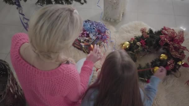 Κάτοψη του ξανθιά ενήλικη γυναίκα και λίγο μελαχρινή κορίτσι κάθεται στο μαλακό χαλί και επιλέγοντας διακοσμήσεις. Μητέρα και κόρη ετοιμάζονται για την Χριστουγεννιάτικη γιορτή. — Αρχείο Βίντεο