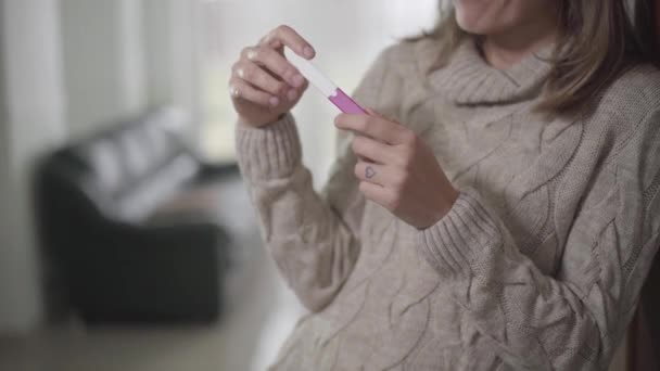 Close-up of young womans hands holding pregnancy test. Blurred Caucasian man passing by at the background and sitting on couch. Wife thinking on the way to tell husband about pregnancy. — Stock Video