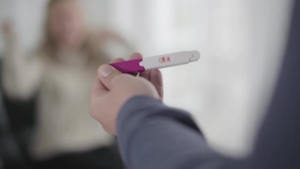 Close-up de mão branca masculina segurando teste de gravidez com duas riscas. Mulher feliz borrada regozijando-se com o fundo. Início de nova vida, gravidez, gestação . — Vídeo de Stock