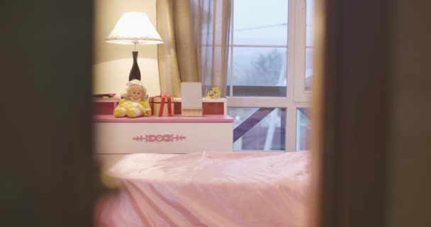 Shooting through doorway of cheerful Caucasian child coming up to pink bedside table, taking doll, and dancing away with the toy. Little girl having fun alone in her room. Leisure, childhood. — Stock Video