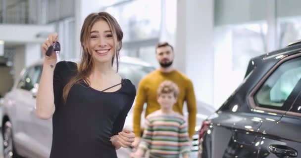 Joven hermosa mujer caucásica jactándose de las llaves del coche y sonriendo. Su marido y su hijo muestran los pulgares hacia arriba en el fondo. Exitosa familia comprando un vehículo nuevo en la sala de exposición. Cinema 4k metraje ProRes HQ . — Vídeos de Stock