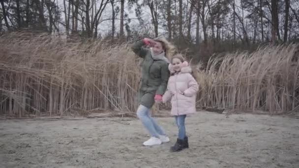 Porträtt av leende vit kvinna som håller handen i pannan och pratar med sin dotter. Glada flicka hoppar och håller mödrar hand. Familjen väntar på någon utomhus på höststranden. — Stockvideo