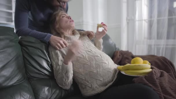 Beautiful pregnant Caucasian blond girl sitting on couch and chewing apple as her husband talking to her and hugging. Young woman getting angry and yelling at man. Pregnancy, gestation, mood swings. — Stock Video