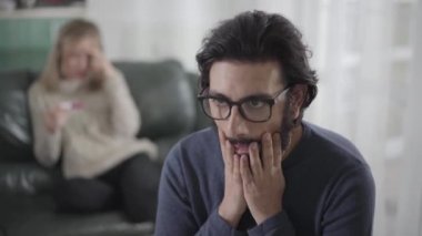 Karısı arka planda hamilelik testiyle koltukta otururken gözlüklü, ön planda sakal süren beyaz bir adam şok oldu. Genç aile gelecekteki bebek için hazır değil. Hamilelik, sorun..