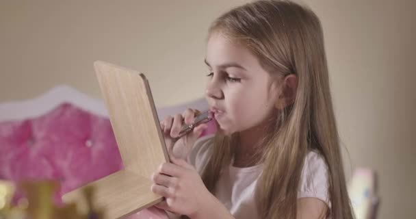 Schattig blank meisje in de slaapkamer met rode lippenstift. Mooi kind dat voor zichzelf zorgt. Gezichtsverzorging, decoratieve cosmetica, kindertijd, make-up. Bioscoop 4k Prores Hq. — Stockvideo