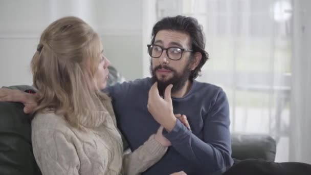 Primo piano di allegra coppia caucasica discutendo mans stile barba. Giovane donna bionda e il suo fidanzato o marito in occhiali relax in casa insieme. Famiglia felice che parla e ride . — Video Stock