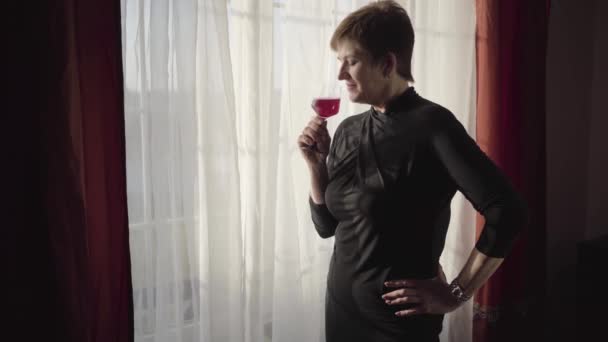 成熟した実業家の香りと赤ワインの試飲。ガラスのワインと窓を眺めながら日光の中に立つスタイリッシュなシニア白人女性の側面図。自信は、休息。レジャー. — ストック動画