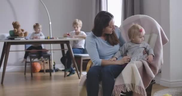 Retrato de mujer caucásica adulta y niña meciéndose en la silla. Feliz madre cuidando de su hija pequeña mientras dos niños más están sentados al fondo. Sede del cine 4k ProRes . — Vídeos de Stock