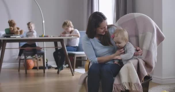 Sonriente mujer caucásica acariciando a la niña y meciéndose en la silla. Feliz madre divirtiéndose con su bebé mientras su hija y su hijo se sientan en el fondo. Sede del cine 4k ProRes . — Vídeos de Stock