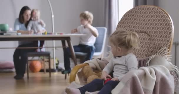 Retrato de una niña rubia caucásica concentrada mirando hacia atrás a su gran familia sentada en el fondo, y jugando con un juguete. Lindo niño sentado en mecedora. Sede del cine 4k ProRes . — Vídeos de Stock