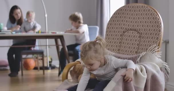 Charmante petite fille blanche soulevant jouet et regardant en arrière sa mère, sa sœur et son frère assis à l'arrière-plan. Portrait d'un joli enfant blond assis sur un fauteuil à bascule. Siège social Cinema 4k ProRes . — Video