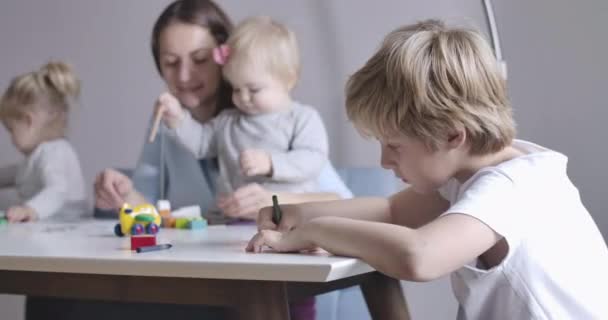 Close-up side view of blond Caucasian schoolboy in white T-shirt drawing. His mother and two sisters sitting at the background and playing. Happy calm day in big family. Cinema 4k ProRes HQ. — Stock Video