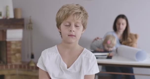 Portrait de garçon caucasien blond aux yeux gris regardant la caméra comme sa sœur et sa mère jouant avec un ours en peluche à l'arrière-plan. Enfant en T-shirt blanc posant à la maison. Siège social Cinema 4k ProRes . — Video