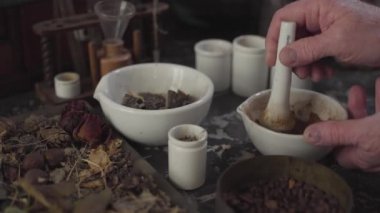 Kurumuş çiçekler ve antika kimyasal konteynırlarla dolu bir masa. Yaşlı beyaz erkek eller havan ve tokmak kullanarak toz öğütüyor. Antik eczacı, alternatif tıp, retro, sağlık.