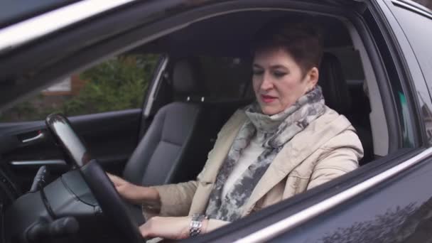 Kısa saçlı, beyaz, olgun bir kadın sürücü koltuğunda oturuyor ve saatine bakıyor. Anne tanınmayan kızını acele etmesi için çağırıyor.. — Stok video