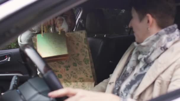 若い白人の女の子は車のサロンで年上の女性にギフトバッグを与えています。母親との娘の出会いと彼女の贈り物を提示。自動車で家族で運転する. — ストック動画