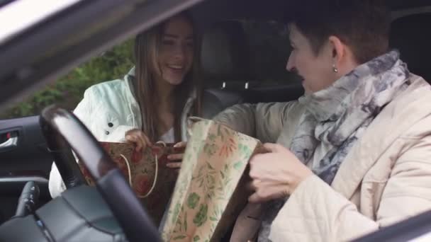 Starsza, dobrze wyglądająca kobieta zagląda do torby z prezentami i przytula córkę. Pozytywna młoda dziewczyna siedząca w salonie samochodowym z mamą i uśmiechnięta. Szczęśliwe rodzinne spotkanie w samochodzie. — Wideo stockowe