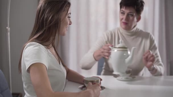 Vista lateral da jovem menina caucasiana sentada à mesa como sua mãe bonita derramando chá em copos. Filha a falar com a mãe e a sorrir. Família passar tempo juntos em casa . — Vídeo de Stock
