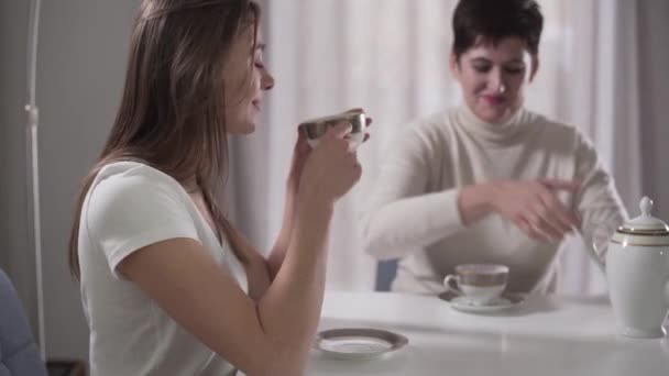 Vue de côté gros plan portrait de jeune fille caucasienne buvant du thé chaud et parlant à la mère assise à l'arrière-plan. Fille profiter du temps avec maman à la maison. Axé sur le premier plan . — Video