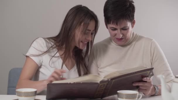 Retrato de dos mujeres caucásicas morenas de diferentes edades viendo álbum de fotos. Madre de aspecto joven y su hermosa hija recordando recuerdos mutuos. Buena relación, vínculo, felicidad . — Vídeo de stock