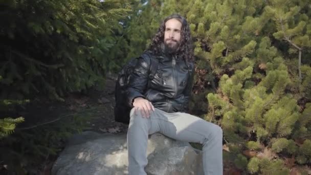 Portrait of confident handsome Middle Eastern man sitting on rock between trees and looking away. Young hippie in leather jacket and jeans resting outdoors. — 图库视频影像