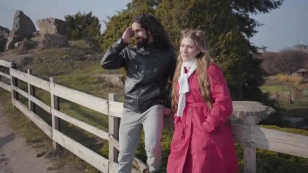 Camera approaching two people standing at wooden fence in autumn park and talking. Relaxed hippie Middle Eastern man and Caucasian woman dating outdoors. Boyfriend and girlfriend resting after walk. — Stock Video