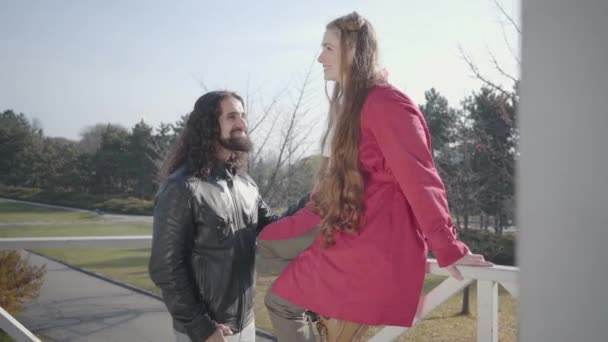 Retrato del hombre barbudo de Oriente Medio con el pelo largo y rizado hablando con su hermosa novia caucásica. Mujer con abrigo rosa sentada en una valla de madera y la cabeza temblorosa. Feliz pareja hippie citas al aire libre — Vídeos de Stock