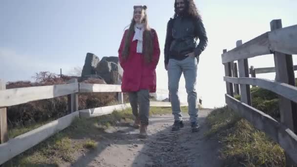 Zwei Menschen laufen die Gasse entlang am Holzzaun entlang. Positives erwachsenes Paar beim Dating im Herbstpark. Hippie-Mann mit langen lockigen Haaren und attraktiver blonder Frau schlendert und redet. — Stockvideo