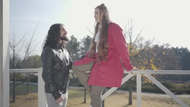 Side view of young hippie man with long black curly hair holding his girlfriend sitting on wooden fence. Happy blond woman talking and smiling. Positive biracial couple dating outdoors. — Stock Video