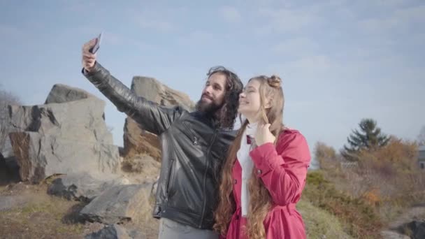 Zijaanzicht van gelukkig Kaukasisch echtpaar trekken gezichten en op zoek naar smartphone scherm. Lachende hippie vriend en vriendin selfie nemen in het najaar park. Positieve man en vrouw daten buiten — Stockvideo
