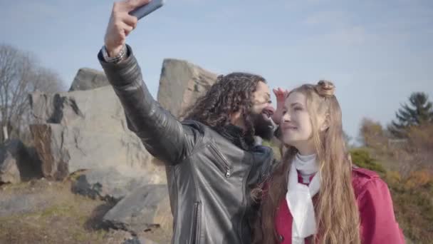 Positiv ung man och kvinna gör selfie i höstparken. Killen med långt lockigt svart hår och blond söt flicka tittar på enheten skärmen, prata och le. Glada hippie par dejtar utomhus. — Stockvideo