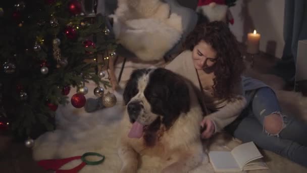 Linda mujer morena caucásica acostada con Moscú Watchdog en alfombra suave y lectura. Chica en ropa casual acariciando y besando a su mascota. Gran perro descansando con su amigo humano en Nochevieja . — Vídeo de stock