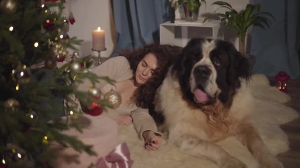 Beautiful adult Caucasian woman lying on big dogs back and sleeping. Saint Bernard guarding his owners dreams on New Years eve. Holidays, celebration, resting, coziness, comfort. — Stock Video
