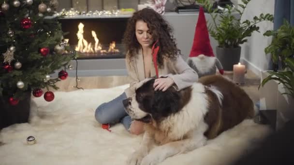 Portrait of pleasant Caucasian girl putting toy horns of deer on dogs head and lying beside him on soft carpet. Beautiful woman decorating Moscow Watchdog on New Years eve. Fun, happiness, joy. — Stock Video
