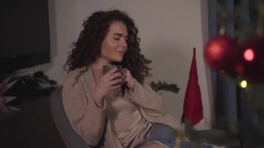 Uzun kıvırcık esmer saçlı güzel beyaz kadının yakın plan portresi evde bir fincan sıcak kahve ya da çayla oturup gülümsüyor. Güzel kız yeni yıl arifesinde dinleniyor. Tatiller, Noel.