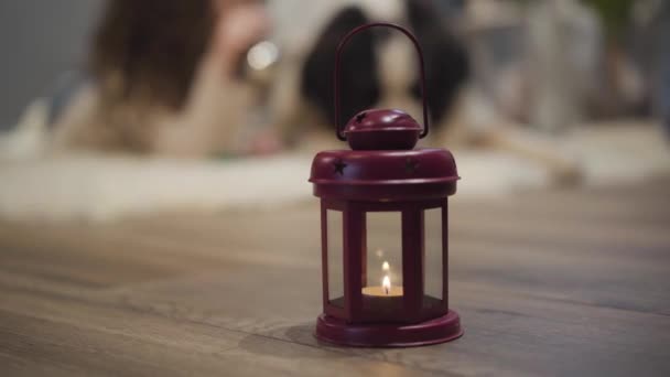 Close-up of New Year candle in lantern. Cheerful blurred Caucasian woman and big dog lying at the background and playing with holiday decoration. Christmas eve, holidays. — Stock Video