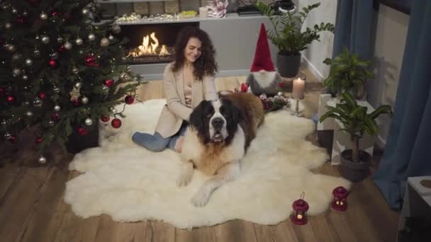 Sorrindo morena caucasiana menina sentada com o cachorro grande no tapete macio na frente da lareira. Árvore de Natal decorada à esquerda. Mulher acariciando seu cão de guarda de Moscou e falando. Feriados . — Vídeo de Stock