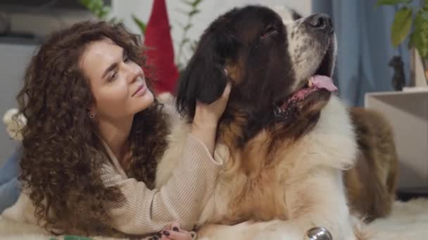 Close-up van grote hond liggend met zijn vrouwelijke eigenaar thuis. Mooie vrouw strelend haar huisdier, kijkend naar de camera en lachend. Vrije tijd, gezelligheid, huisdieren. — Stockvideo
