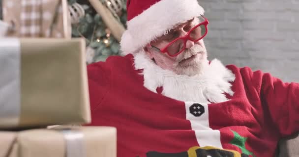 Close-up portret van uitgeputte Kerstman slapen als zittend op de bank met geschenkdozen. Oude blanke man met witte baard met rode hoed, rustend op oudejaarsavond. Bioscoop 4k Prores Hq. — Stockvideo