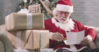 Konsantre olmuş yaşlı Noel Baba 'nın mektup okuma portresi. Gözlüklü, beyaz sakallı, çocuk için hediye seçen olgun bir adam. Arka planda duran süslü Noel ağacı. Sinema 4k Prores Merkezi.
