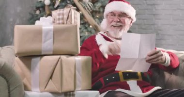 Beyaz sakallı Noel Baba 'nın portresi mektup okuyor, hediye kutularına bakıyor ve gülümsüyor. Gözlüklü son sınıf öğrencisi yeni yıl arifesinde çocuk için hediye seçiyor. Sinema 4k Prores Merkezi.