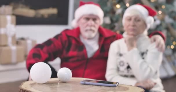 Mettre l'accent sur les changements de smartphone pour les visages de triste couple âgé assis à côté de l'arbre de Noël. Les retraités attendent l'appel des enfants la veille du Nouvel An. Siège social Cinema 4k ProRes . — Video