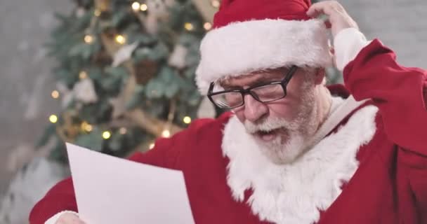 Zbliżenie zmęczonego Świętego Mikołaja w okularach, drapiącego się po głowie jak list do czytania. Staruszek myśli o prezencie dla dziecka w Sylwestra. Kino 4k Prores Hq. — Wideo stockowe