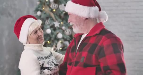 Vrolijk stel dat voor de kerstboom danst. Glimlachende oude man en vrouw met rode hoeden die oudejaarsavond samen thuis doorbrengen. Bioscoop 4k Prores Hq. — Stockvideo